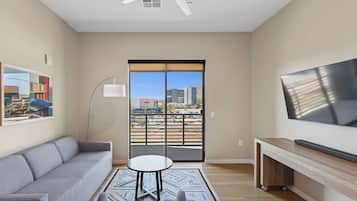 Una sala de estar moderna con un sofá gris, una mesa de centro redonda y un televisor de pantalla plana montado en la pared. Se ve una ventana grande con vistas a edificios y un balcón.