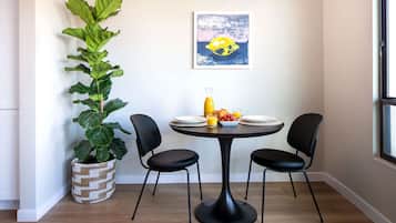 A dining area with a round table, black chairs, a potted plant, a fruit bowl, and a framed artwork on the wall.