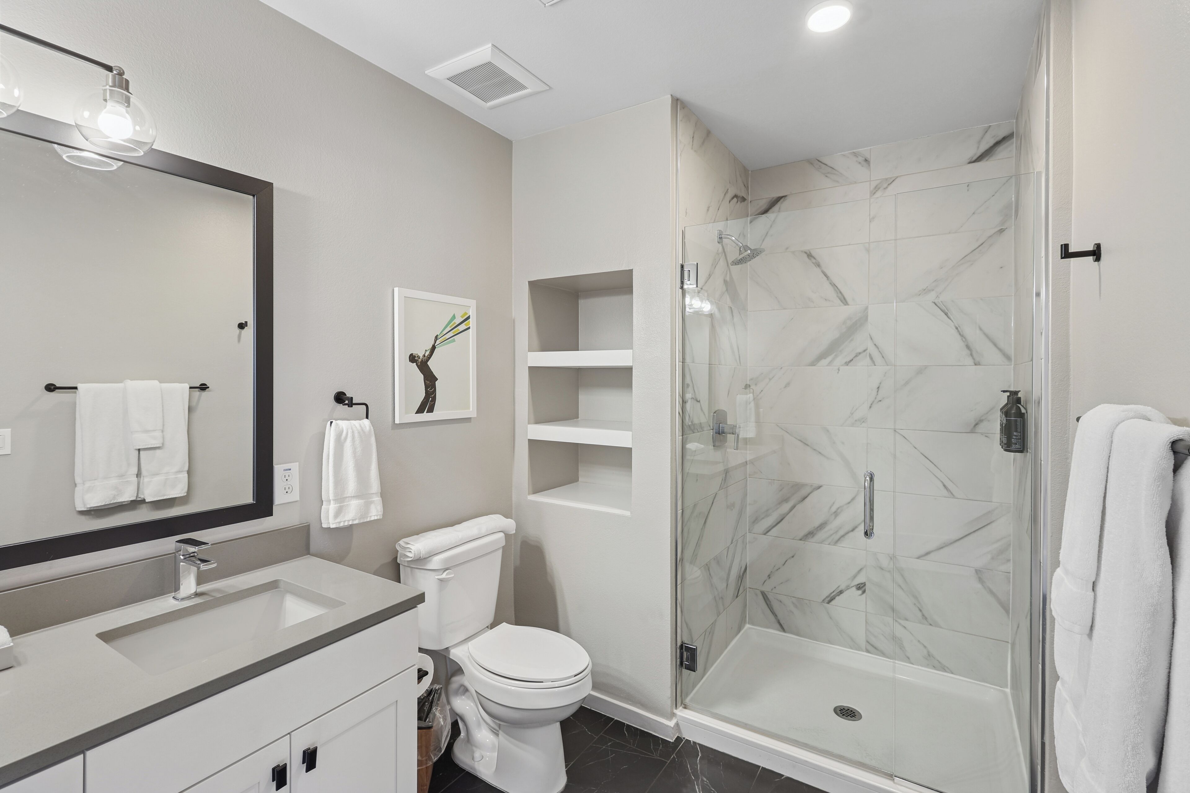 A modern bathroom with a large mirror, white fixtures, a glass-enclosed shower, and marble tiles.