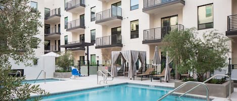 Un immeuble d’appartements moderne doté d’une piscine, d’un espace extérieur aménagé et d’une zone couverte avec des parasols.