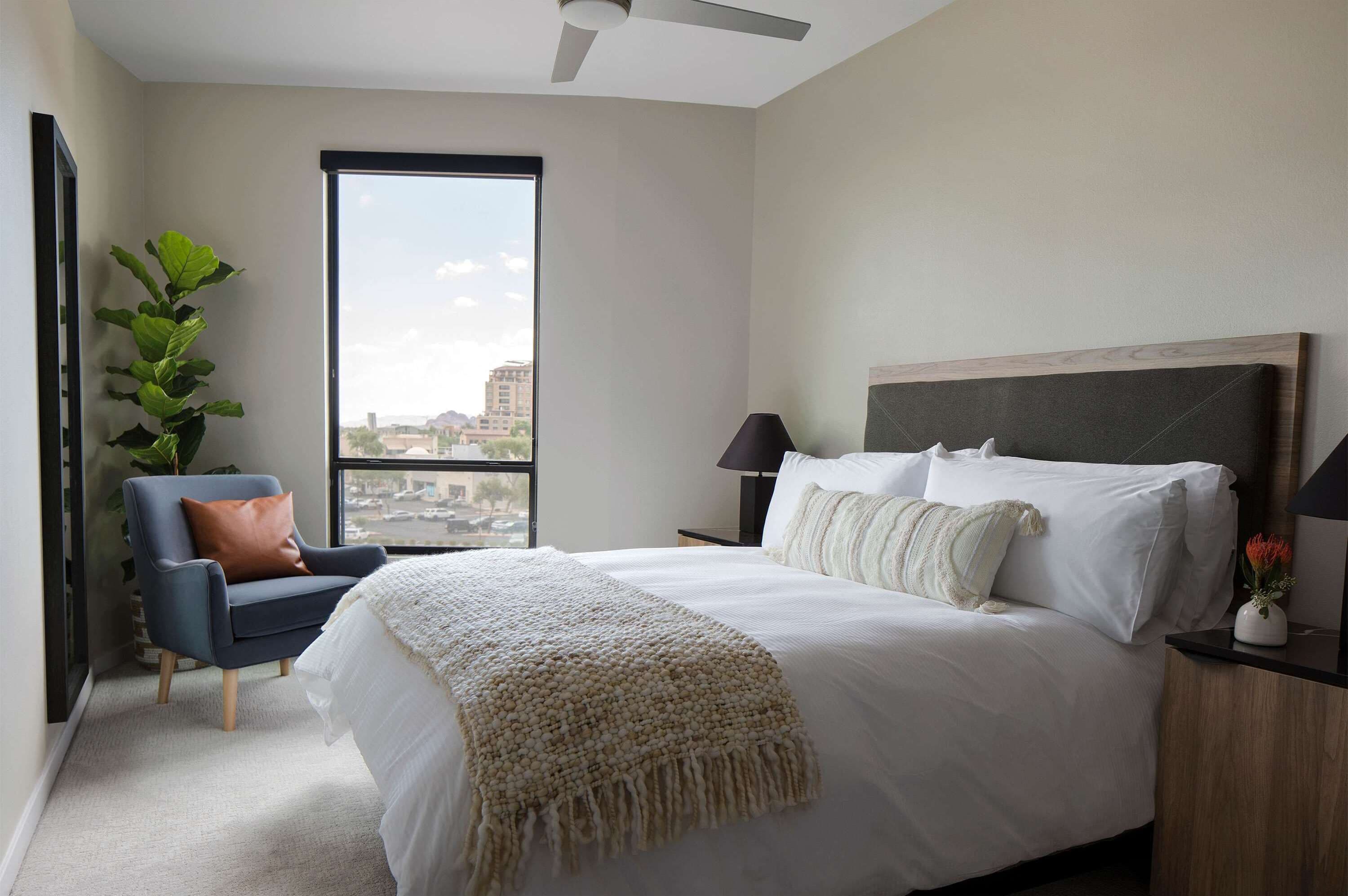 A bedroom with a large bed, a chair, a nightstand, a lamp, a plant, and a view of the city through a window.