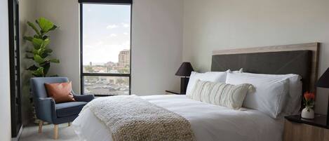 A bedroom with a large bed, a chair, a nightstand, a lamp, a plant, and a view of the city through a window.