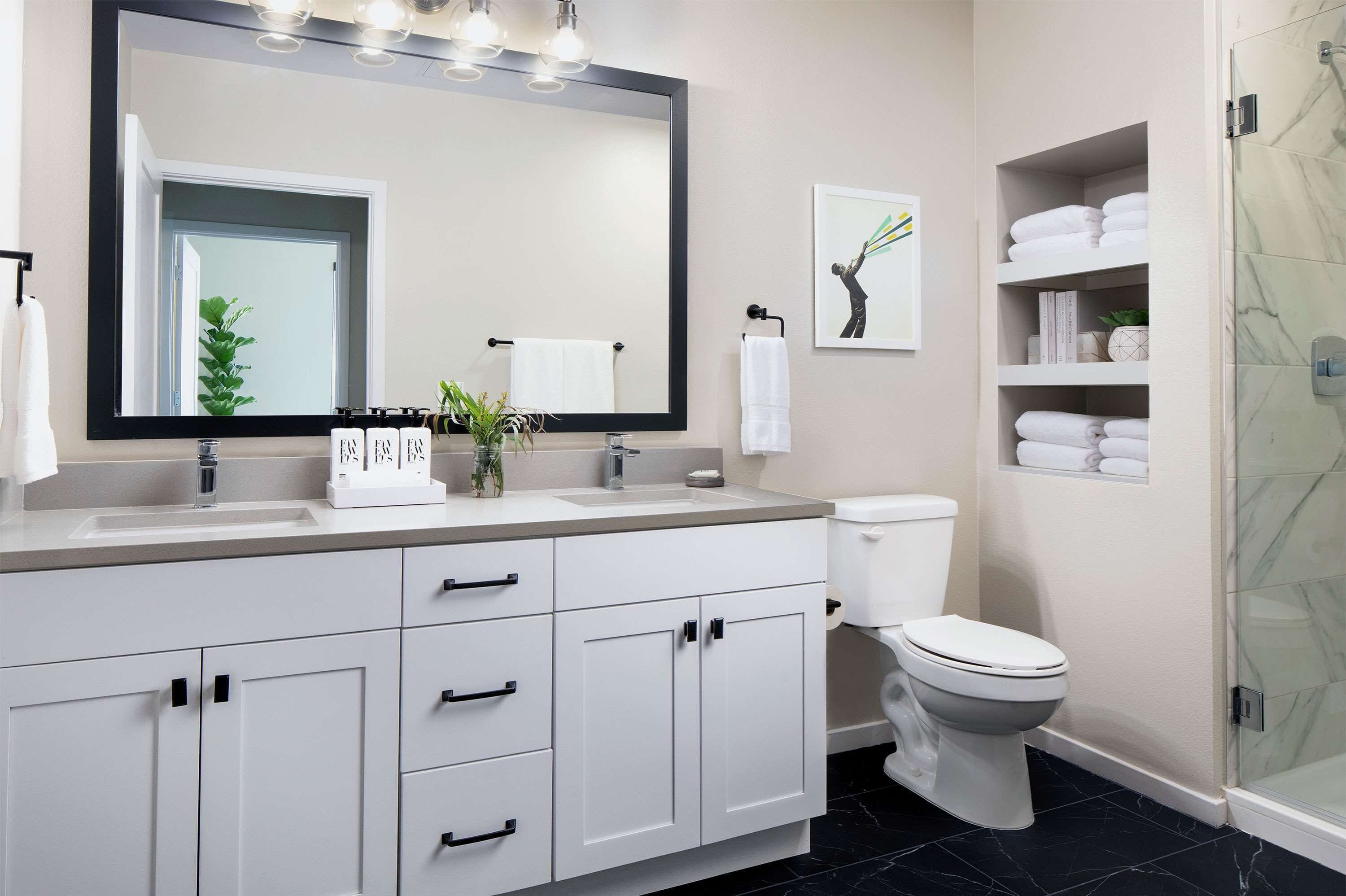 A modern bathroom with a large mirror, two sinks, a toilet, and a glass shower enclosure.