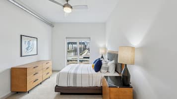 A bedroom with a bed, a dresser, a nightstand, a ceiling fan, and a window with a city view.