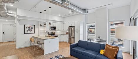 Un apartamento moderno con cocina, comedor y sala de estar. Cuenta con electrodomésticos de acero inoxidable, un sofá azul y una mesa de centro redonda.