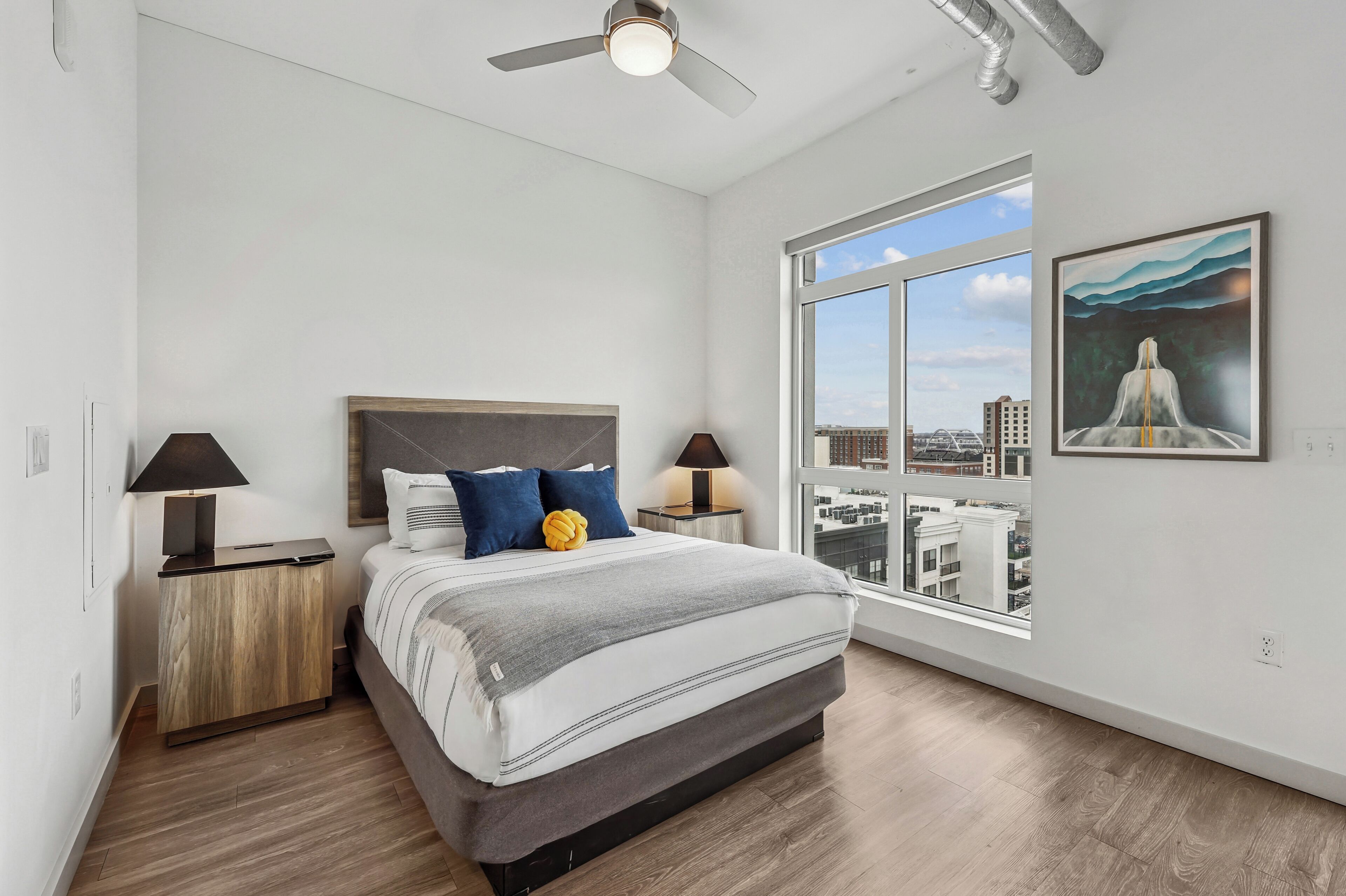 Un dormitorio moderno con una cama grande, dos mesitas de noche con lámparas, un ventilador de techo y un ventanal con vistas a la ciudad.
