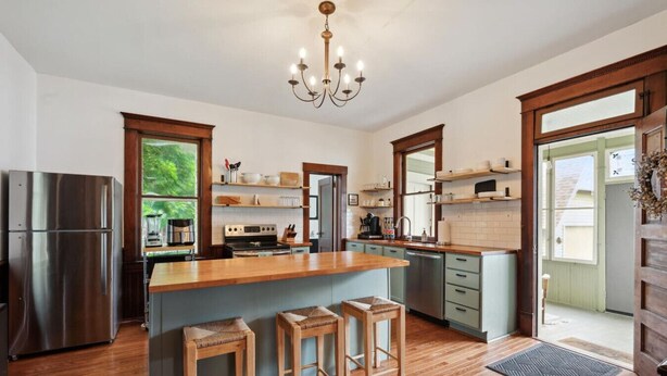 A modern kitchen with a central island, wooden stools, a stainless steel refrigerator, and a chandelier.