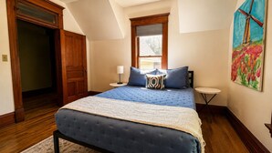 A bedroom with a large bed, a window, a door, and a painting on the wall. - Cloverleaf Victorian | Historic Victorian | Walk Downtown | Sleeps 10 (Pella)