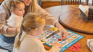 A family playing a board game at a wooden table in a room with a window and a chair.