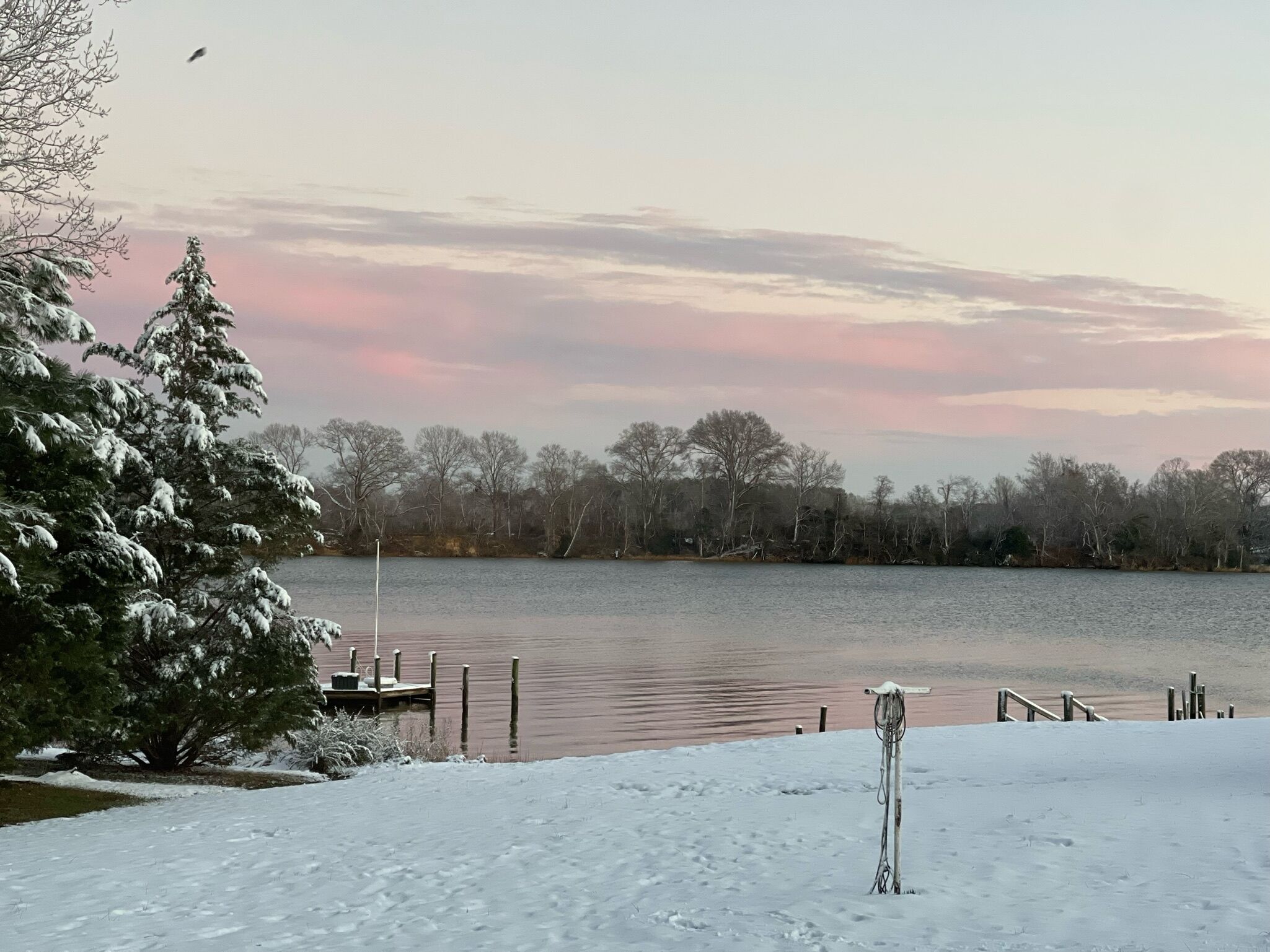 A snow-covered landscape with a lake, dock, and trees.
