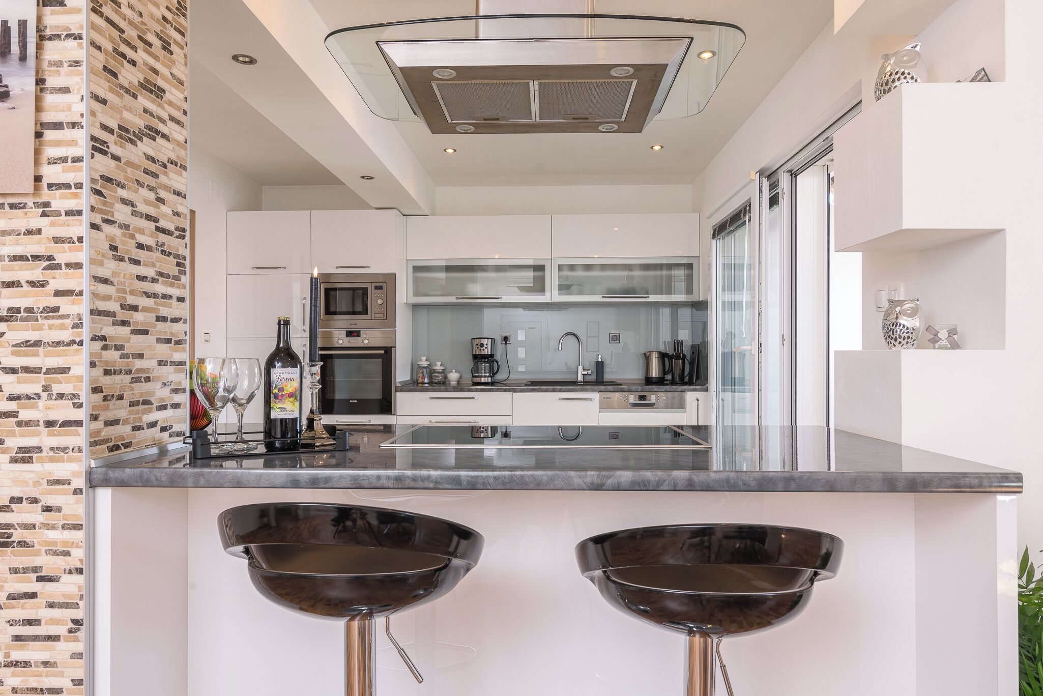 A modern kitchen with white cabinets, a stainless steel refrigerator, and a glass backsplash. There are two bar stools with metal bases and no seats.