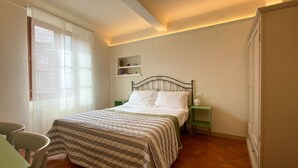 Un dormitorio con cama, una silla, una mesita de noche y un estante con libros.