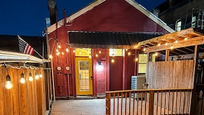A wooden deck with a wooden fence, a wooden bench, and a pathway leading to a red building with a yellow door. - Main Street 2BR Apartment with Garage and Deck (Saint Charles)