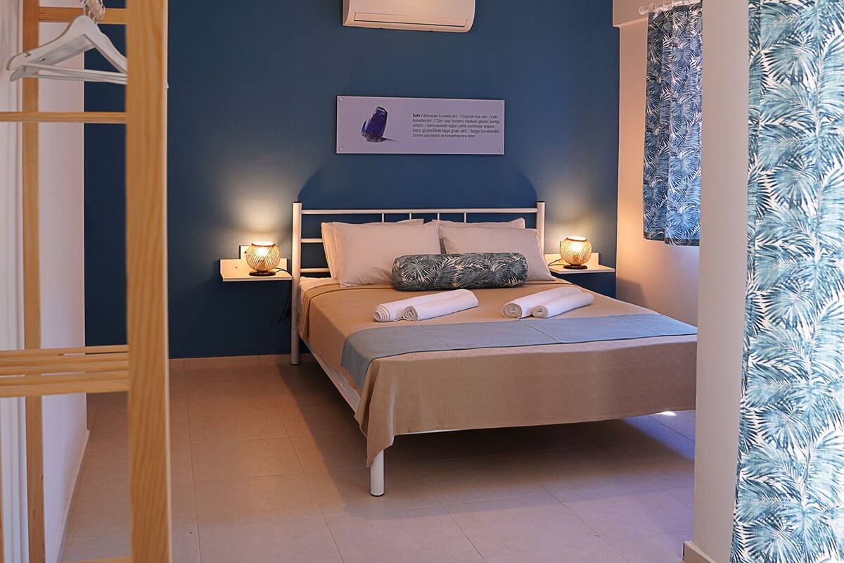 A bedroom with a bed, two bedside tables, a hanging light fixture, a wall-mounted air conditioner, and a window with a curtain.