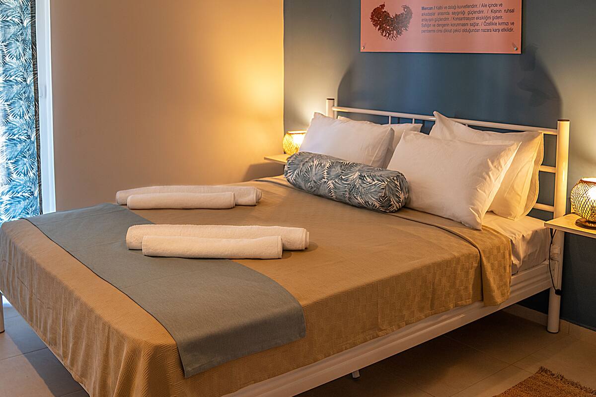 A neatly made bed with a brown bedspread and two rolled towels, a patterned pillow, and a wall-mounted sign with text.