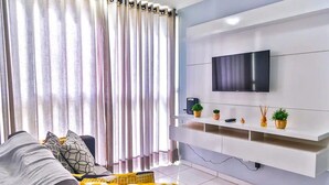 A modern living room with a sofa, a flat-screen TV mounted on a white wall, and a shelf with decorative items. - Ap Centro no Coração de BC - 500m do Mar (Balneário Camboriú)