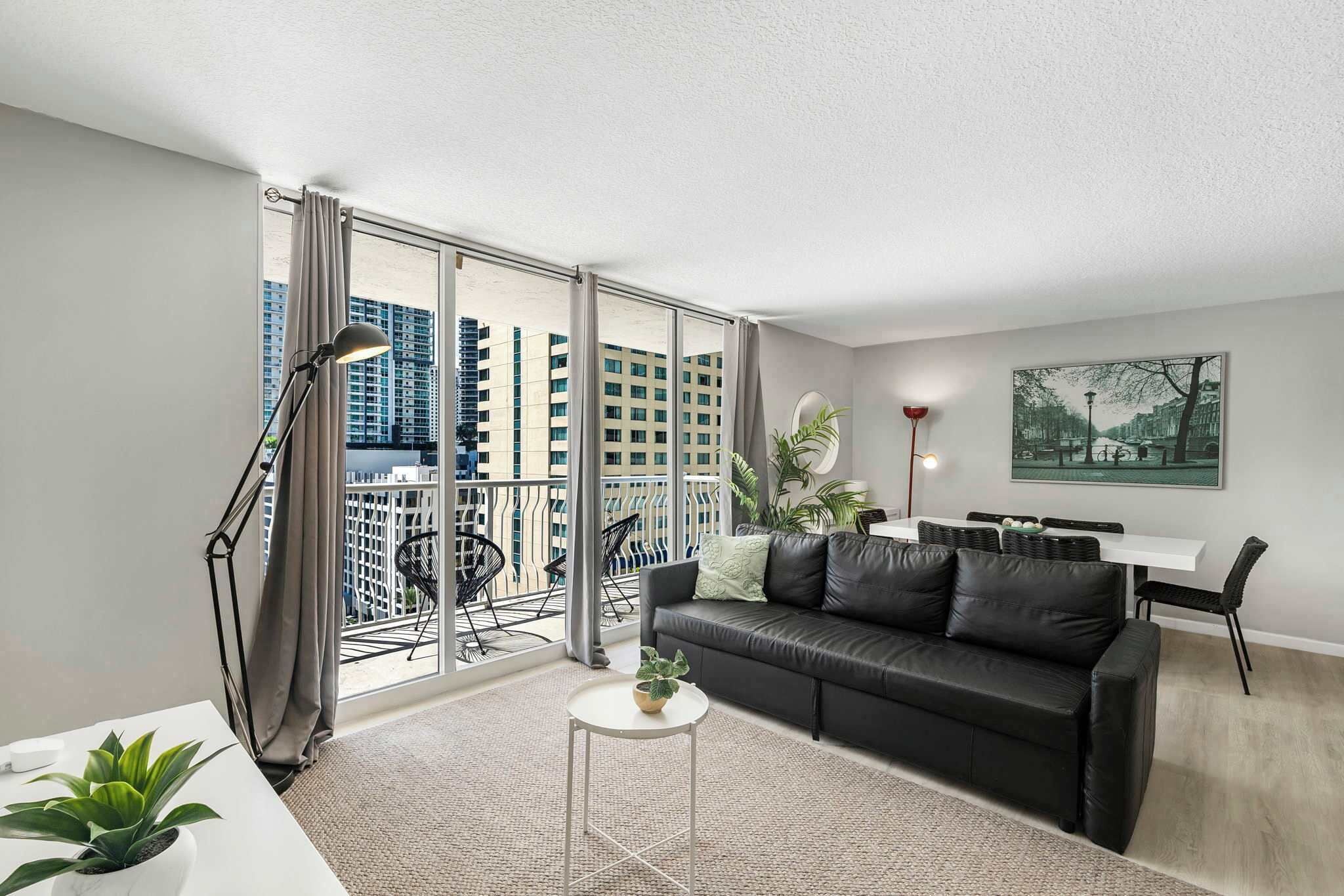 A modern living room with a black leather sofa, a small white table, and a balcony with a view of high-rise buildings.