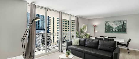 A modern living room with a black leather sofa, a small white table, and a balcony with a view of high-rise buildings.