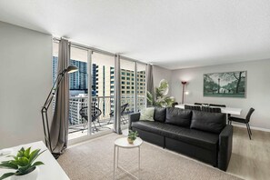 Un salon moderne avec un canapé en cuir noir, une petite table blanche et un balcon offrant une vue sur les immeubles de grande hauteur.