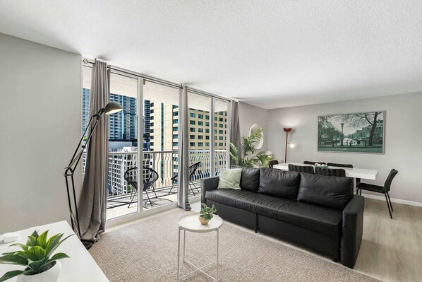 A modern living room with a black leather sofa, a small white table, and a balcony with a view of high-rise buildings.