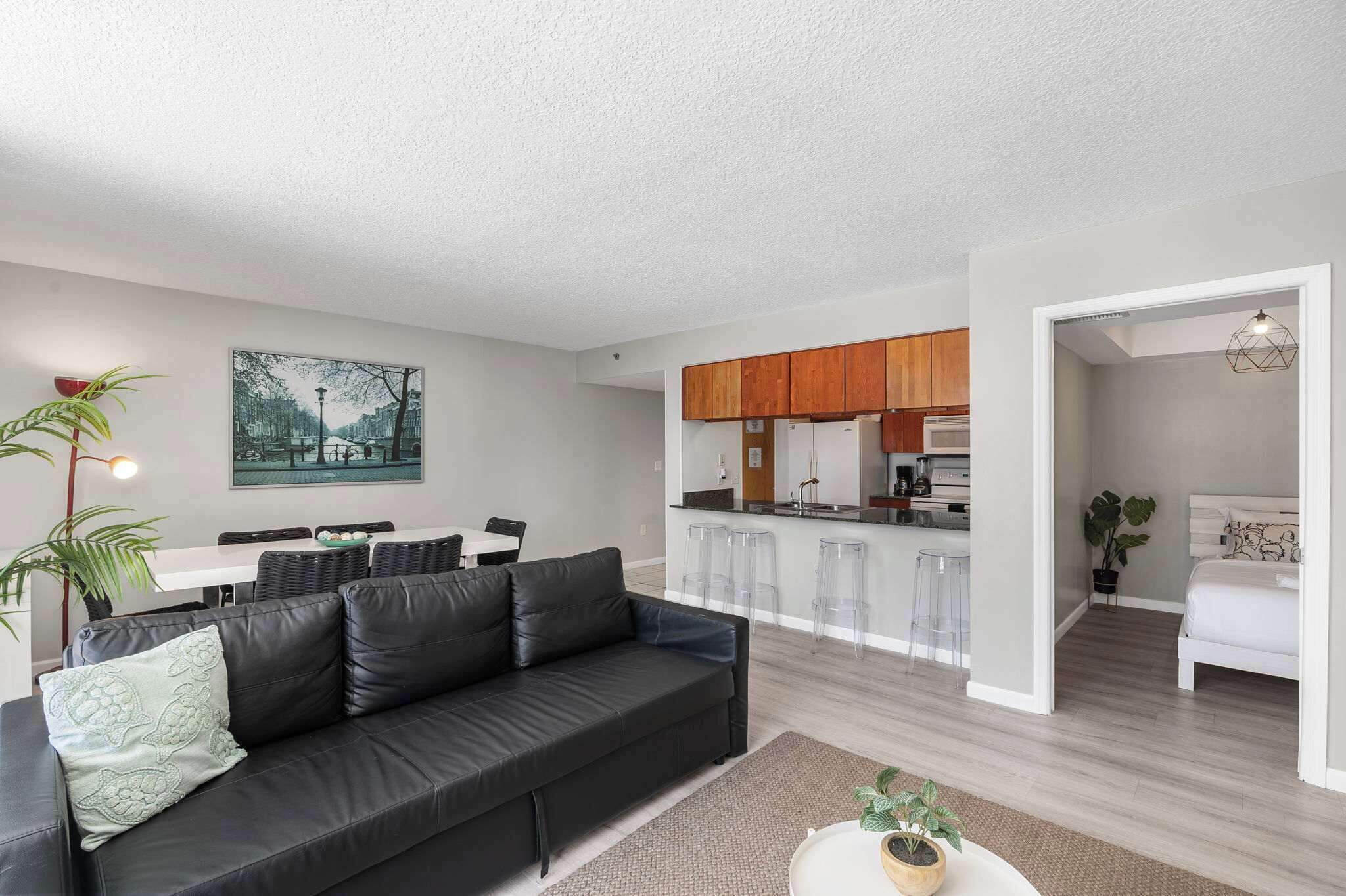A modern living room with a black leather sofa, a dining area with a white table, and a kitchen with wooden cabinets.