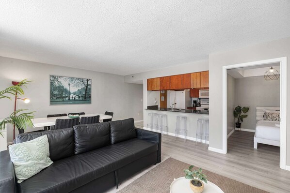 A modern living room with a black leather sofa, a dining area with a white table, and a kitchen with wooden cabinets.