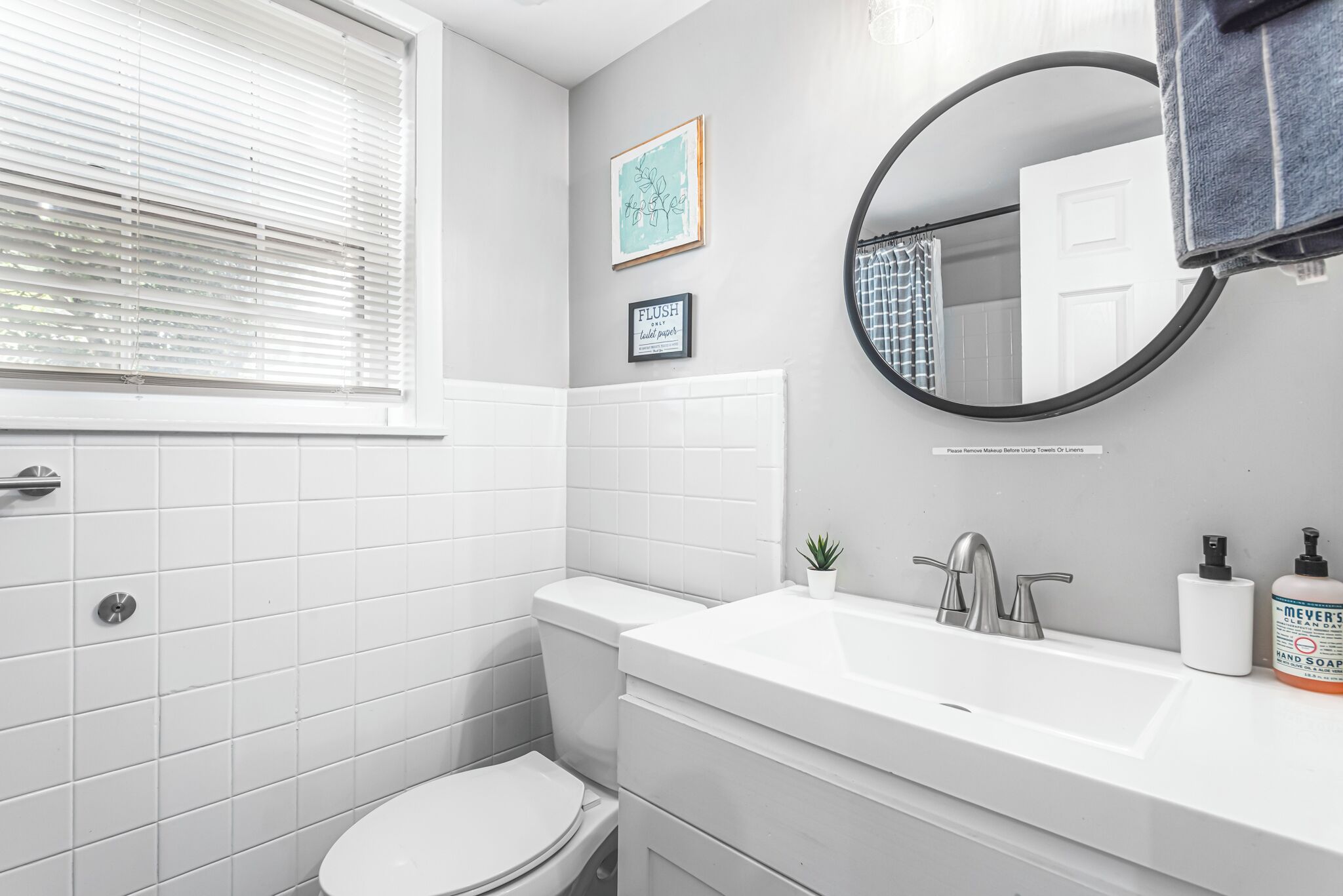 Une salle de bain avec des carreaux blancs, des toilettes, un lavabo avec un miroir et une fenêtre avec des stores.
