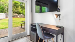 A room with a sliding glass door leading to a backyard, a desk with a chair, a mounted TV, and a desk lamp.
