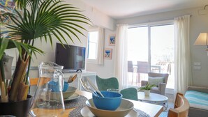 A well-lit living room with a television, dining table, chairs, and a large window with curtains. - Carreta Sea Views by Hello Homes Sitges (Sitges)