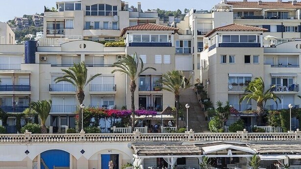A beachfront hotel with multiple balconies, palm trees, and a beach area with outdoor seating. - Apartment by the Sea by Hello Homes Sitges (Sitges)