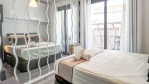 A modern hotel room with a large bed, a sitting area with a chair, a small table, and a large window with a view of the city. - Flamingo Apartment by Hello Homes Sitges (Sitges)