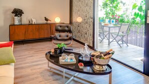 A modern living room with a leather armchair, a coffee table with a bottle of champagne, glasses, and a bowl of strawberries. A wooden cabinet with decorative items and a bird sculpture is visible. - Flamingo Apartment by Hello Homes Sitges (Sitges)