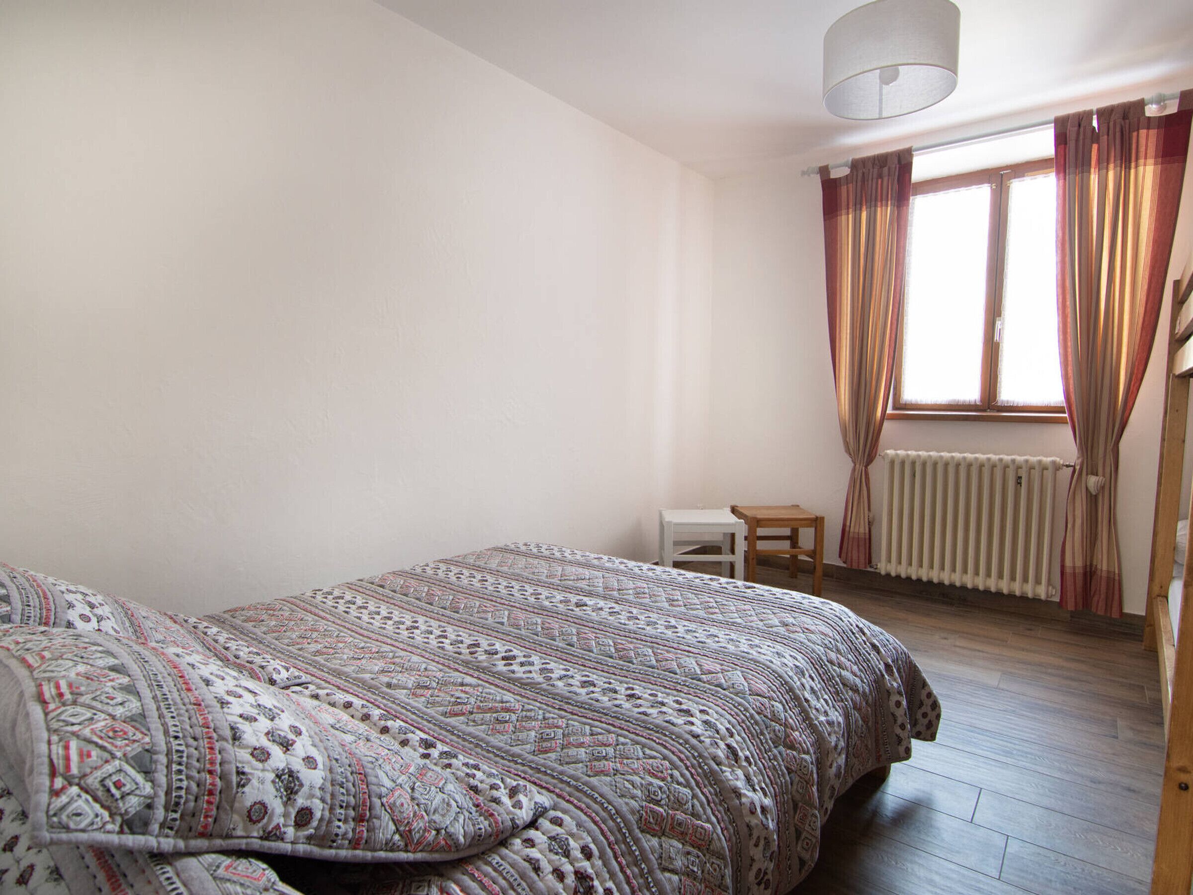 A bedroom with a bed, a small table, a window with curtains, and a radiator.