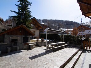 A stone patio with a wooden table and benches, surrounded by wooden buildings with a mountain backdrop. - 3-room Southeast-facing Apartment for 6-8 People with Wifi (Valloire)