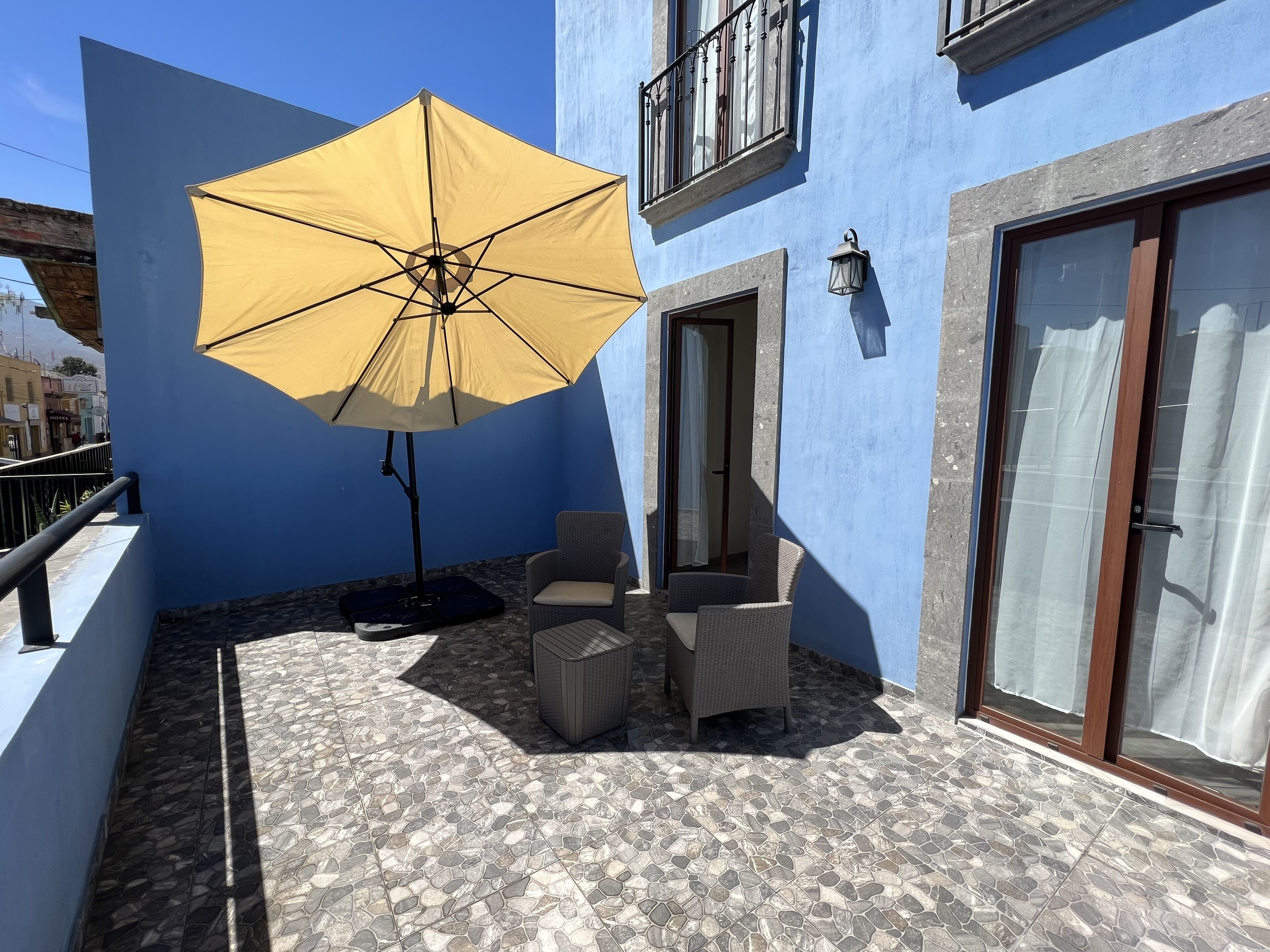 Uma varanda com um guarda-sol amarelo, duas cadeiras de vime, uma pequena mesa e um chão de azulejos.