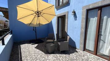 Uma varanda com um guarda-sol amarelo, duas cadeiras de vime, uma pequena mesa e um chão de azulejos.