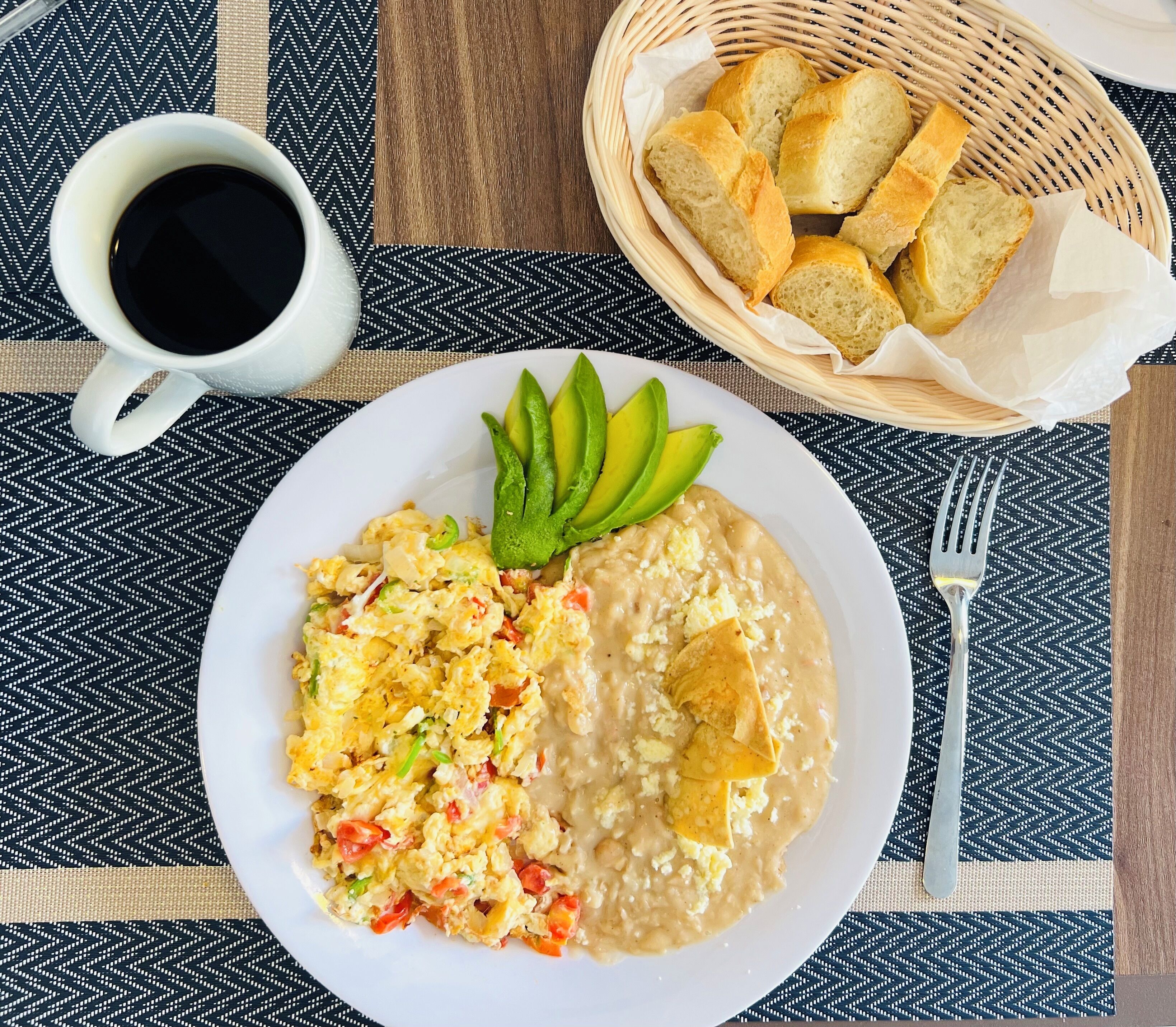 A plate with scrambled eggs, refried beans, and avocado slices, accompanied by a cup of black coffee and a basket of bread.