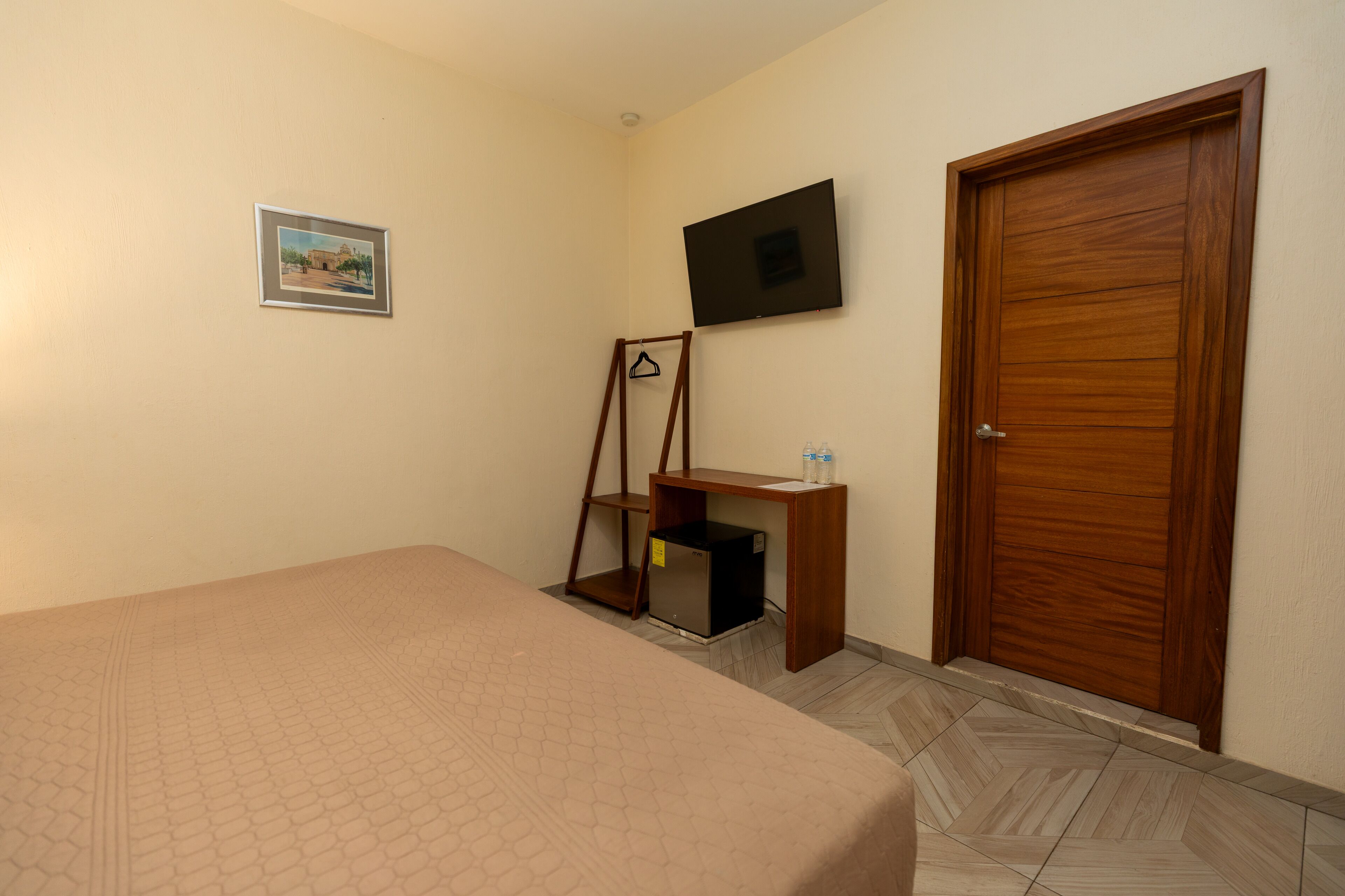 Un dormitorio con cama, televisor montado en la pared, puerta de madera y un escritorio pequeño con estante.