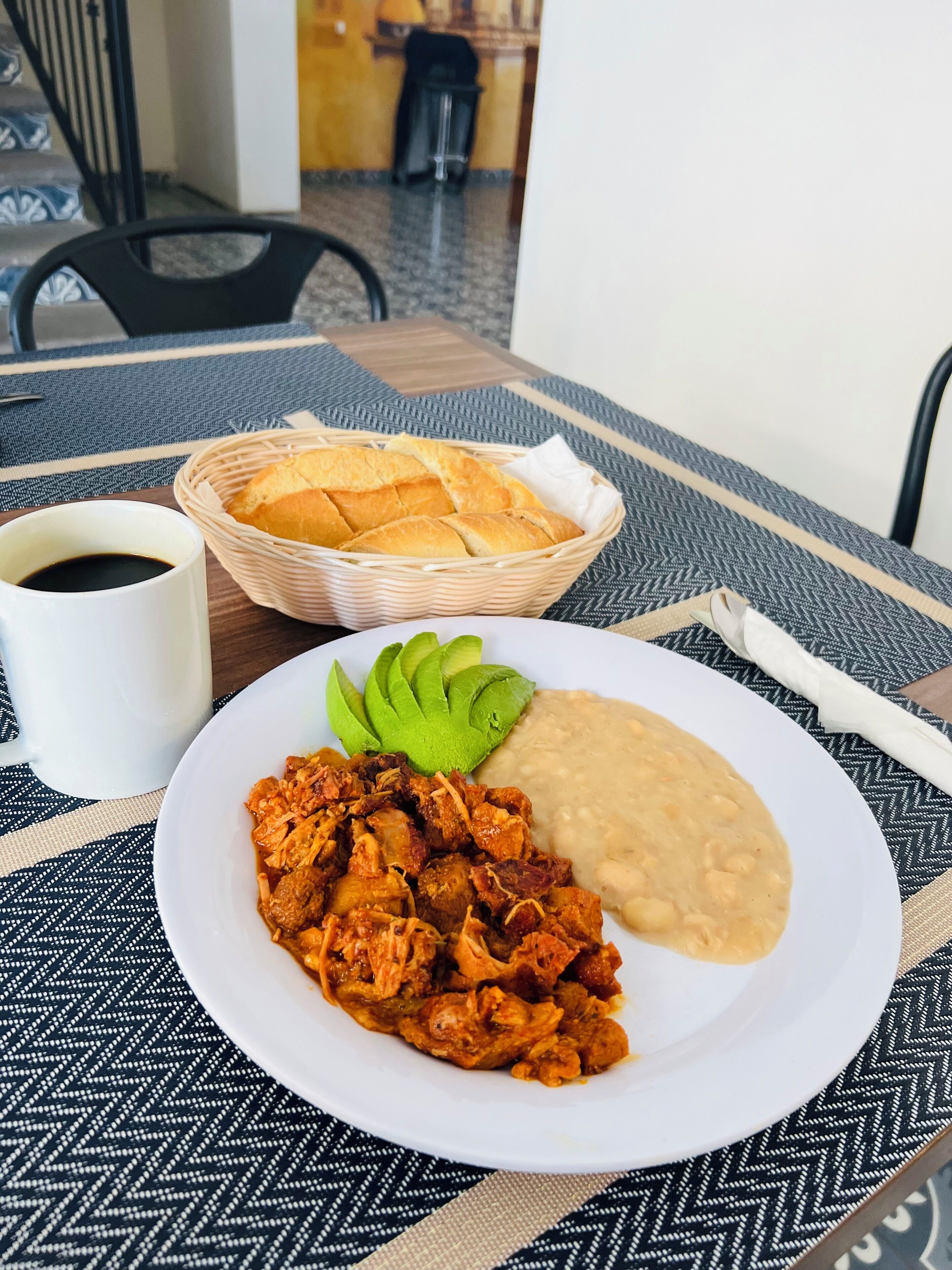 Une assiette de nourriture comprenant des tranches d’avocat, des haricots et un plat de viande, un panier de pain et une tasse de café, le tout posé sur une table à motifs.