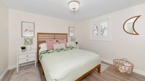 A bedroom with a wooden bed, white nightstands, a decorative ceiling light, framed artwork, and a window with blinds. - The Ivanhoe Getaway (Sarasota)