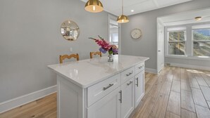 Una cocina moderna con una isla blanca, sillas de madera, luces colgantes, un jarrón con flores, un espejo redondo y un reloj en la pared.