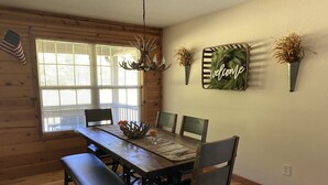 A wooden dining room with a large table, benches, and chairs. There is a chandelier, a window with blinds, and a welcome sign on the wall. - Riverfront Cabin! Mountain Views, Pool, Hot Tub, Fishing, Tubing, Fire Pit, WIFI (Hiawassee)