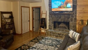 A living room with a stone fireplace, a television mounted above it, a brown leather couch, a patterned rug, and a wooden shelf with decorative items. - Riverfront Cabin! Mountain Views, Pool, Hot Tub, Fishing, Tubing, Fire Pit, WIFI (Hiawassee)