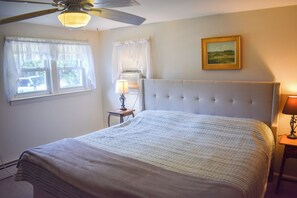 Un dormitorio con una cama grande, un ventilador de techo, una ventana con cortinas, una mesita de noche con una lámpara y un cuadro en la pared.
