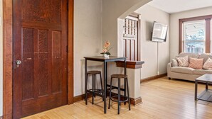 Una habitación con puerta de madera, una mesa pequeña con taburetes, un sofá y un televisor montado en la pared.