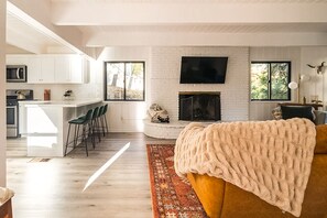 A modern living room with a fireplace, a kitchen area with white cabinets, and a dining space with bar stools.