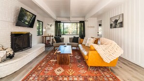 A living room with a fireplace, a sofa, a coffee table, and a TV mounted on the wall. - Family & Pet-Friendly, Cozy & Stylish Cabin Steps from Lake+Village (Lake Arrowhead)