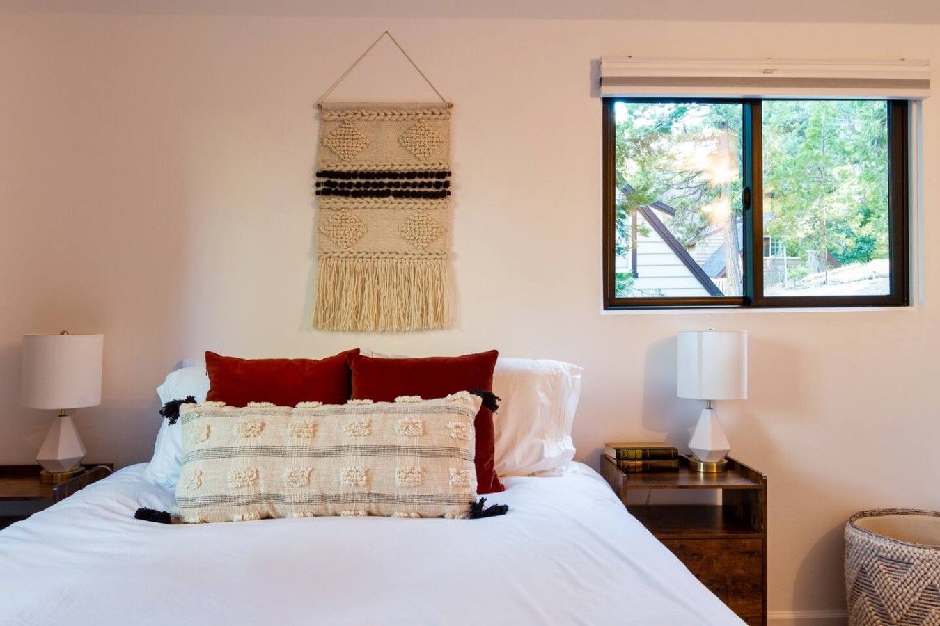 A bedroom with a bed, two bedside lamps, a wall hanging, and a window with a view of trees.
