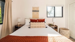 A bedroom with a large bed, two bedside tables, a hanging macrame wall hanging, a window with a view, and a waste basket. - Family & Pet-Friendly, Cozy & Stylish Cabin Steps from Lake+Village (Lake Arrowhead)