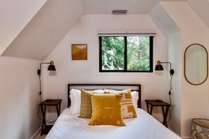 Un dormitorio con cama, dos mesitas de noche, ventana con vista a árboles, espejo en la pared y un marco de fotos.